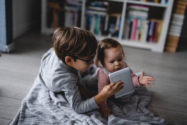 Küçük beyaz kız bebek evde yerde sürünürken abisi dijital tablet erkek ve kız kardeşinin çocukluğunu gösteriyor. Tablet ve ele odaklı seçici bir konsept olarak yetişiyor.