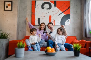 Bir grup Kafkasyalı aile üyesi, anne ve baba ve çocukları evde kanepede oturmuş oyun konsolu ile oyun oynuyorlar. Oyun çubuğu ya da kumandayla mutlu kazanmalar ve gülümsemeler.