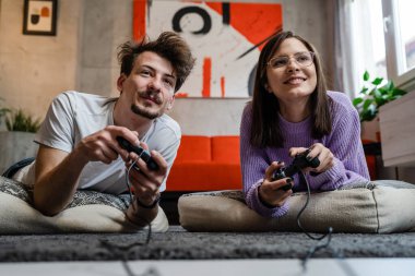 Kadın ve erkek evlerinde yerde yatıp oyun konsolu oynuyorlar. Eğlenceli genç çiftler birlikte zaman geçiriyorlar. Oyun kumandası tutuyorlar.