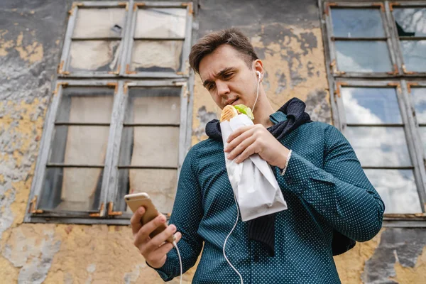 携帯電話を使用して音楽を聴いたり 明るい日に建物の外でサンドイッチを保持し 食べている間に呼び出しを行うために1人の若い白人男性大人現実の人々高速食品コピースペース — ストック写真