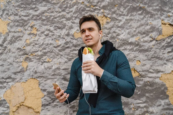 携帯電話を使用して音楽を聴いたり 明るい日に建物の外でサンドイッチを保持し 食べている間に呼び出しを行うために1人の若い白人男性大人現実の人々高速食品コピースペース — ストック写真