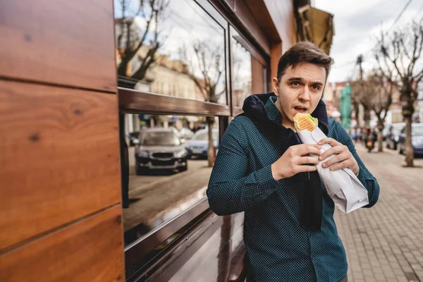 秋の春や冬の日に通りに立っている男が 足で食べるジャケットを着たサンドイッチを持ち 建物の前で屋外で待っている現実の人々はスペースをコピーしますファーストフードの概念 — ストック写真