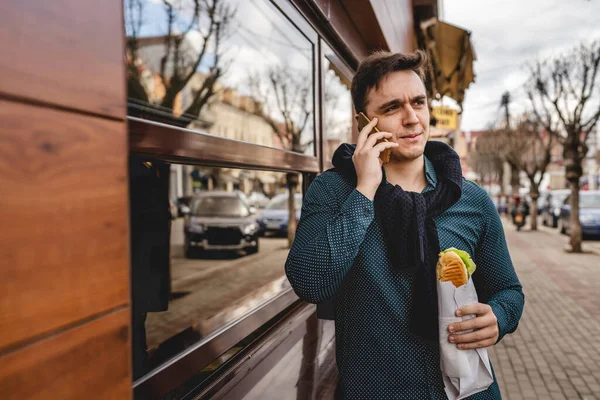 明るい日の建物の横にサンドイッチの屋外を保持し 食べている間に通話を作るために携帯電話を使用して1人の若い白人男性大人高速食品コピースペース — ストック写真