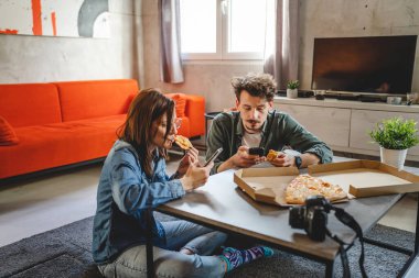 İki kişi, güzel bir çift, beyaz bir erkek ve kadın, evde oturmuş pizza yiyor ve cep telefonlarını sosyal ağ için kullanıyor. İnternette geziniyorlar, birlikte oturuyorlardı.