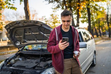 Genç bir beyaz adam kaputu açık ve bozuk motorlu aracının yanında duruyor. Sonbahar günü yol kenarında yardım için çekici çağırıyor.
