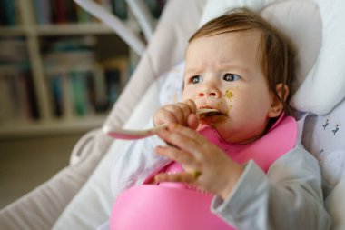 Küçük bir Kafkas bebek zekası yüzünü kirletiyor. Ağzına ve çenesine yiyecek doluyor. Elinde plastik kaşıkla salıncakta bekliyor.