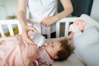 Beyaz kadın annenin, yeni doğmuş bebeğine baktığı iki aylık şişenin süt ya da mama ile beslendiği ve çocuğun beşikte seçici odak noktasında yattığı görüntüsü.