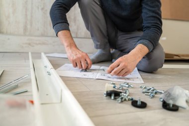 Kimliği belirsiz beyaz bir erkek evde yerde oturuyor. Eşyaları birleştiriyor. Elinde parçalar ve talimatlar tutuyor.