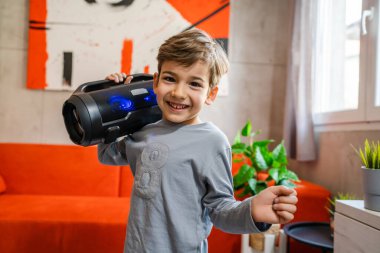 Küçük mutlu bir beyaz çocuk elinde kablosuz ağ hoparlörü ile evde müzik dinliyor.