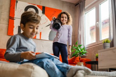 Çocuklar evde tek başlarına eğlenirken iki kardeş ellerinde wifi hoparlörüyle müzik dinlerken ağabeyi dijital tablet kullanarak evde eğleniyor.