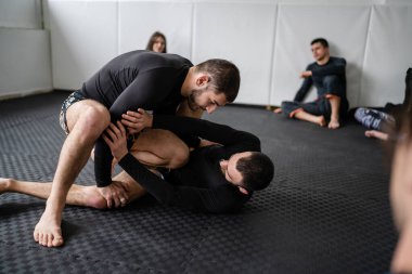 two men demonstrate bjj brazilian jiu jitsu grappling or luta livre technique on the ground at training at the academy in front of group of students leg attack