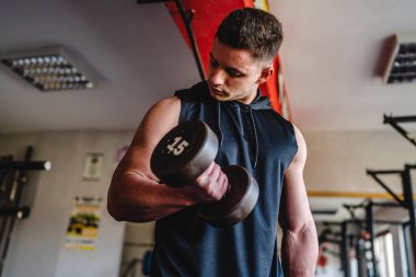 Yetişkin, beyaz, erkek, vücut geliştiren, kollarını çalıştıran, spor salonunda dikilirken kaslarını halterle esneten bir adam.