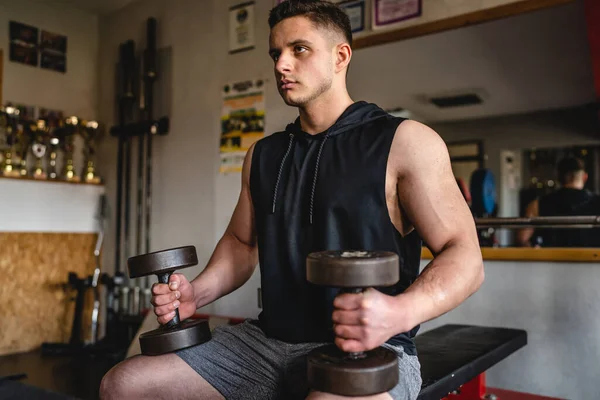 一位年轻的男性健美运动员穿着衬衫坐在体育馆里 一边用哑铃训练双臂二头肌弯曲肌肉 一边模仿空间前视镜腰部 — 图库照片