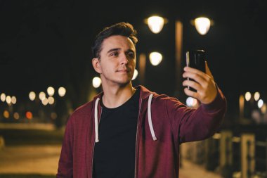 Genç, beyaz bir adamın portresi gece veya gündüz şehir veya kasabada cep telefonuyla video görüşmesi ya da selfie fotokopi çekilen uzay belini kaldırırken çekilmiş.