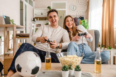 İki beyaz insan evlerinde oturup video oyunu konsolu oynayarak eğleniyorlar. Genç çift, erkek ve kız arkadaş birlikte vakit geçiriyorlar. Oyun kumandası ellerinde gülümsüyorlar.