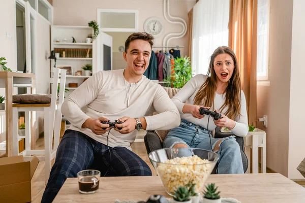 İki beyaz insan evlerinde oturup video oyunu konsolu oynayarak eğleniyorlar. Genç çift, erkek ve kız arkadaş birlikte vakit geçiriyorlar. Oyun kumandası ellerinde gülümsüyorlar.
