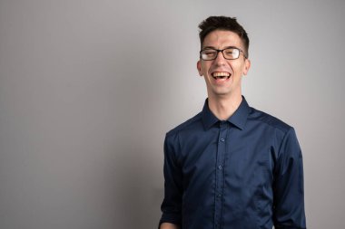 25 yaşında beyaz gözlüklü yetişkin bir adamın portresi. Beyaz duvarın önünde kameraya bakıyor. Sırıtıyor.