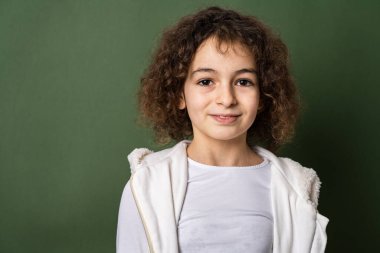 On yaşında, kıvırcık saçlı, yeşil arka planda dikilip kameraya bakıp gülümseyen mutlu ve neşeli fotokopi çeken bir beyaz kız.