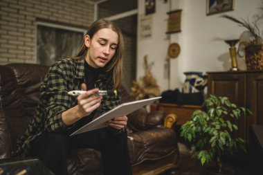 Beyaz tenli genç bir kadın sanatçı evinde, kanepede oturup resim yapıyor elinde boya fırçası ve yaratıcılık, profesyonel ya da hobi kavramları var.