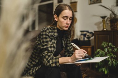 Beyaz tenli genç bir kadın sanatçı evinde, kanepede oturup resim yapıyor elinde boya fırçası ve yaratıcılık, profesyonel veya hobi kavramı var.