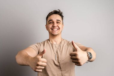 Beyaz duvarın önünde sıradan kıyafetler giyen genç bir beyaz adam olumlu jest yapıyor. Gülümsüyor ve mutlu bir yüz ifadesi sergiliyor. Mutlu bir gülümseme.