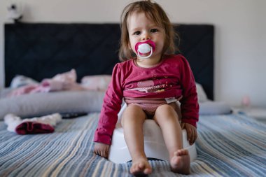 Bir yaşında beyaz bir kız bebek. Evde, çocuk tuvaletinde gün boyu yatağında oturuyor. Meme ucu emziği ağzında.