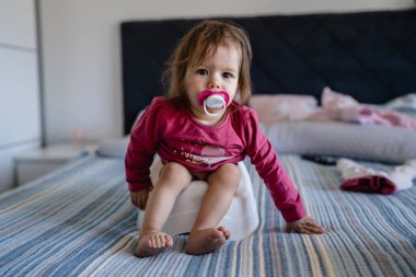 Bir yaşında beyaz bir kız bebek. Evde, çocuk tuvaletinde gün boyu yatağında oturuyor. Meme ucu emziği ağzında.