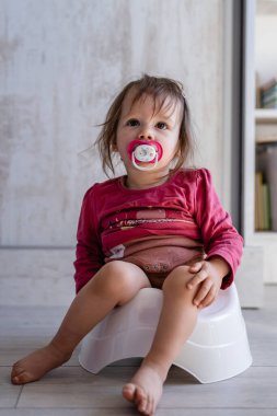 Bir yaşında küçük beyaz bir kız bebek. Evde, çocukların tuvaletinde oturuyor. Gün boyu ağzında meme ucu emziği, fotokopi odası.