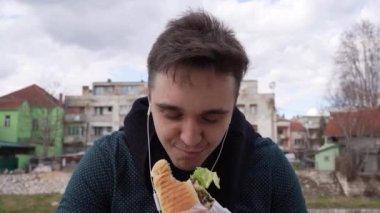 Genç beyaz bir erkek, şehirde ya da şehirde açık havada sandviç yiyor ve tutuyor. Gerçek insanlar fast food kavramını ön plana çıkarıyor. Yavaş çekim kapanıyor.