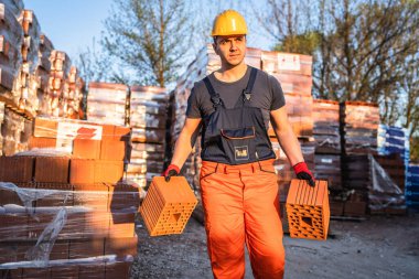 Bir inşaat işçisi, güneşli yaz gününde kil portakalı blokları depo ya da şantiye alanlarını alıp taşıyor ya da tutuyor.