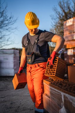 Bir inşaat işçisi, güneşli yaz gününde kil portakalı blokları depo ya da şantiye alanlarını alıp taşıyor ya da tutuyor.