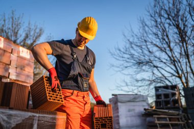 Bir inşaat işçisi, güneşli yaz gününde kil portakalı blokları depo ya da şantiye alanlarını alıp taşıyor ya da tutuyor.
