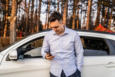 Genç, yetişkin bir işadamı, arabasının önünde dikilirken cep telefonu kullanıyor. Gömlek giyiyor, meşgul görünüyor. Gerçek insanlar yer kopyalıyor.