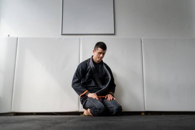 Akademi erkek sporcusu Brezilya jiu jitsu dersi BJJ jiujitsu konsepti için hazırlanıyor. Tatami minderlerinin üzerinde tek başına oturuyor. Kimono gi üniforması giyiyor.