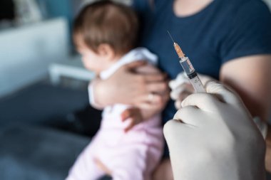Kimliği belirsiz doktor koruyucu eldivenler giyip elinde şırıngayla küçük beyaz anne için aşı hazırlarken bebeğini aşı için kucağında tutuyor.