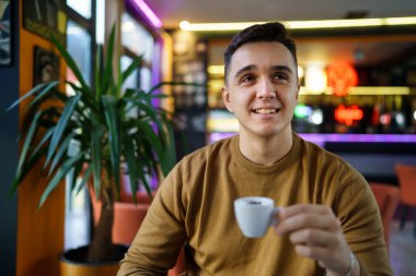 Kafeteryada tek başına oturan beyaz bir adam elinde bir fincan kahve fotokopi alanı tutan mutlu bir erkeği görüyor.