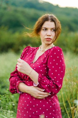 Yazın yeşil tarlada dikilen güzel, doğal görünümlü beyaz bir kadının yarım boy portresi. Kırmızı elbiseyle manken pozu veriyor.