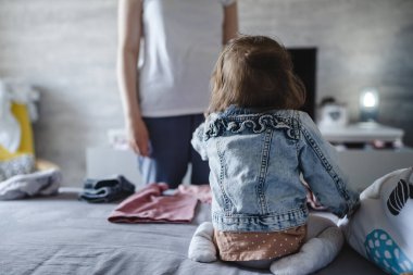 Back view of small caucasian baby girl sitting on the bed at home watching her mother taking care of her real people family childhood growing up and care concept copy space