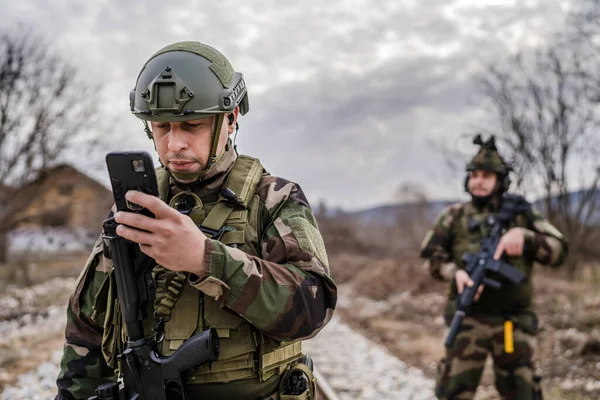Two soldiers dogs of war mercenaries men in uniform armed service rifles standing while securing the railroad in combat zone in war soldier use mobile phone while patrolling area during mission