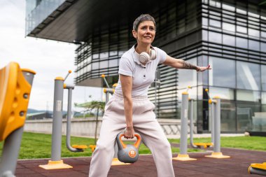 Gündüz bina önünde kısa saçlı, modern, olgun bir beyaz kadın spor salonunda ağırlık sallıyor sağlıklı yaşam tarzı konseptini taklit ediyor.