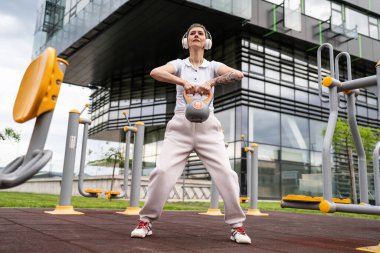 Bir kadın, modern olgun, beyaz kadın. Kısa saçlı, bina önünde günün ilerleyen saatlerinde spor salonunda kettlebell ağırlığı ile spor salonu sağlıklı yaşam tarzı konseptini taklit ediyor.