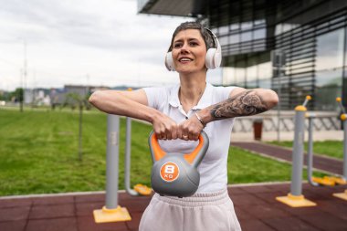 Bir kadın, modern olgun, beyaz kadın. Kısa saçlı, bina önünde günün ilerleyen saatlerinde spor salonunda kettlebell ağırlığı ile spor salonu sağlıklı yaşam tarzı konseptini taklit ediyor.