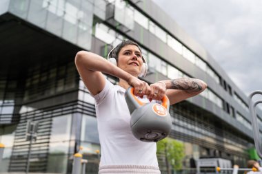 Bir kadın, modern olgun, beyaz kadın. Kısa saçlı, bina önünde günün ilerleyen saatlerinde spor salonunda kettlebell ağırlığı ile spor salonu sağlıklı yaşam tarzı konseptini taklit ediyor.
