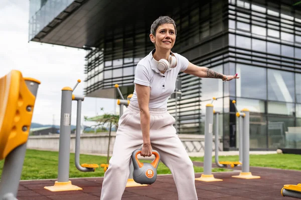 Gündüz bina önünde kısa saçlı, modern, olgun bir beyaz kadın spor salonunda ağırlık sallıyor sağlıklı yaşam tarzı konseptini taklit ediyor.