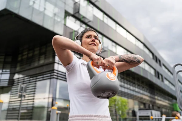Bir kadın, modern olgun, beyaz kadın. Kısa saçlı, bina önünde günün ilerleyen saatlerinde spor salonunda kettlebell ağırlığı ile spor salonu sağlıklı yaşam tarzı konseptini taklit ediyor.