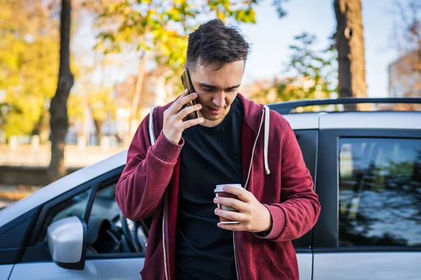 町のコピースペースで路上で待っている間側に見てテイクアウトコーヒーのカップを保持している秋の日や夜に彼の車のそばに立つ若い白人男性電話を話して作る — ストック写真