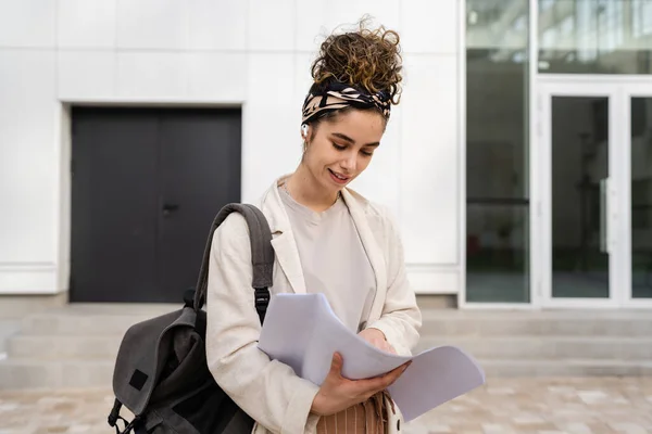 一个年轻的女商人或女学生在明亮的日子里在大学或公司的现代建筑前查阅文件 真实的人独自站着抄写空间手握着纸快乐的微笑 — 图库照片