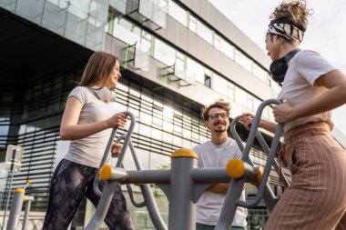 Bir grup arkadaş, erkek ve kadın, erkek ve genç, açık havada spor salonunda, modern binanın önünde antrenman yapıyorlar. Gerçek insanlar spor ve eğlence, sağlıklı yaşam tarzı kavramları geliştiriyorlar.