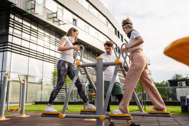 Bir grup arkadaş, erkek ve kadın, erkek ve genç, açık havada spor salonunda, modern binanın önünde antrenman yapıyorlar. Gerçek insanlar spor ve eğlence, sağlıklı yaşam tarzı kavramları geliştiriyorlar.