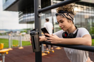 Beyaz bir kadın açık hava antrenmanı sırasında fren yapıyor açık hava spor salonunda barlarda dinleniyor el sıkışma aleti ile su içiyor ya da ek olarak mutlu bir gülümseme fotokopisi çekiyor.